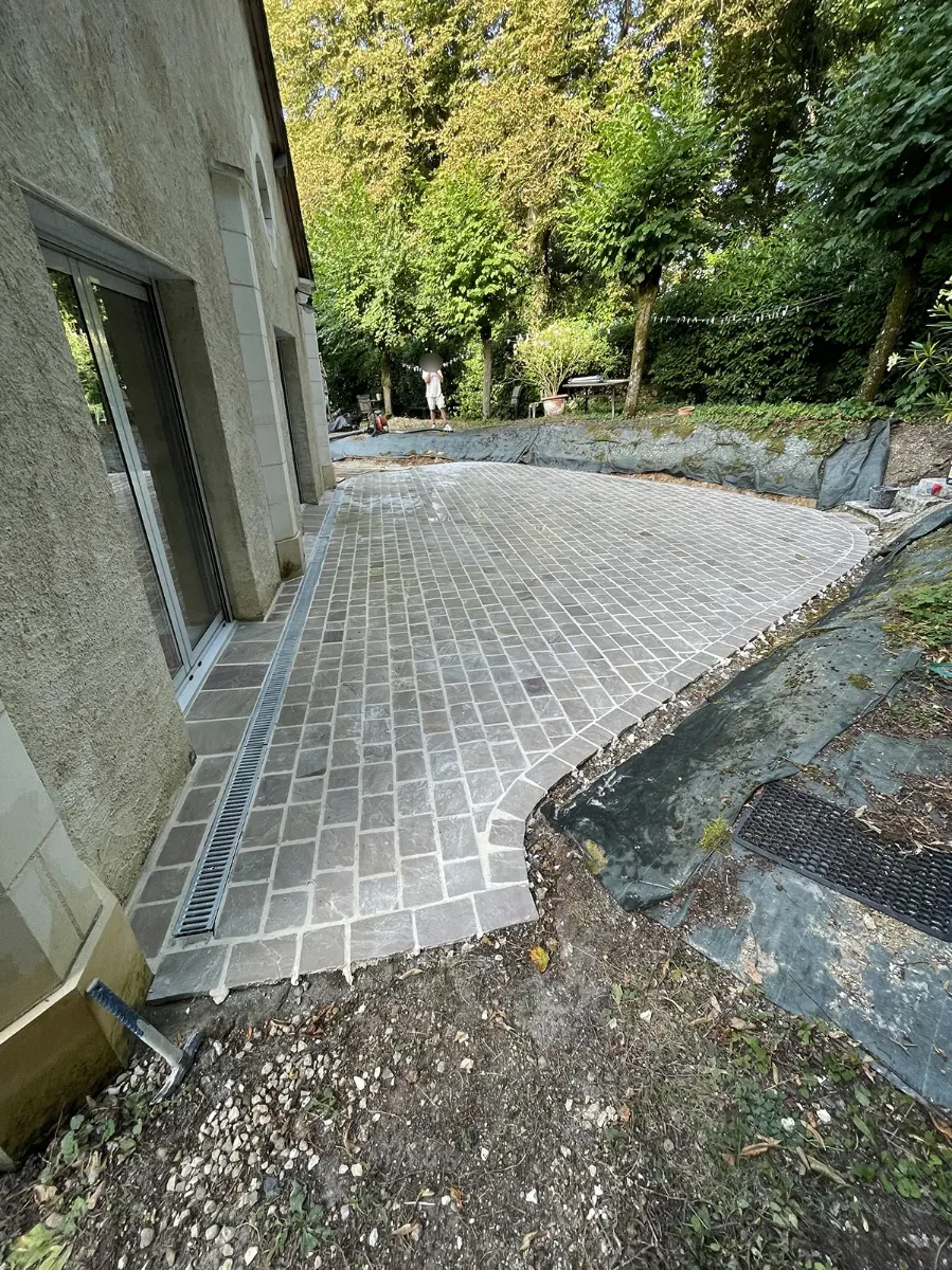 Terrasse en pavés de pierre achevée le long d'une maison, caniveau de façade et talus bâché