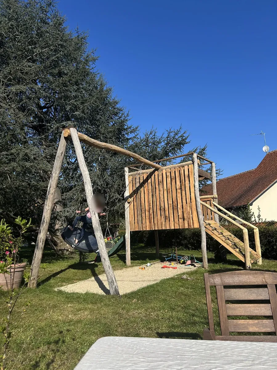 Aire de jeux en bois avec cabane sur pilotis, escalier, toboggan et balançoire nid, sous un ciel bleu