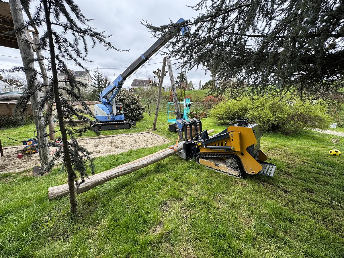Grue araignée et chargeuse compacte transportant un poteau de bois sur le chantier d'une aire de jeux