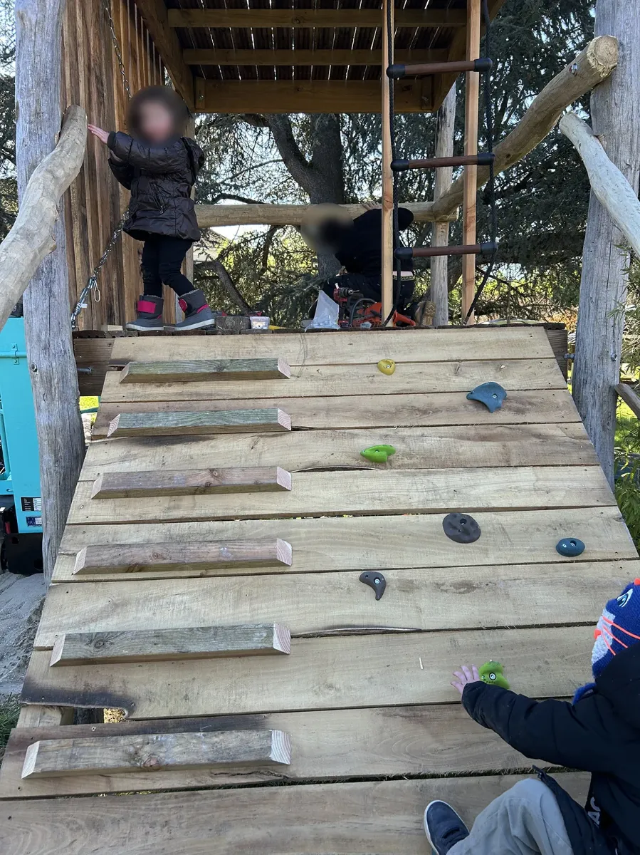 Rampe d'escalade en planches équipée de prises colorées, menant à la plateforme d'une cabane de jeux