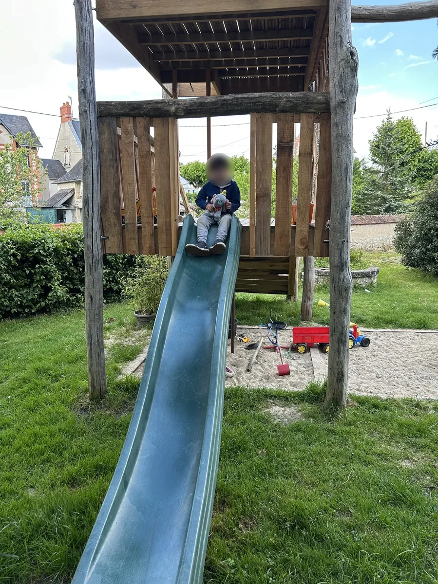 Toboggan vert descendant de la plateforme d'une cabane de jeux vers un bac à sable, dans un jardin