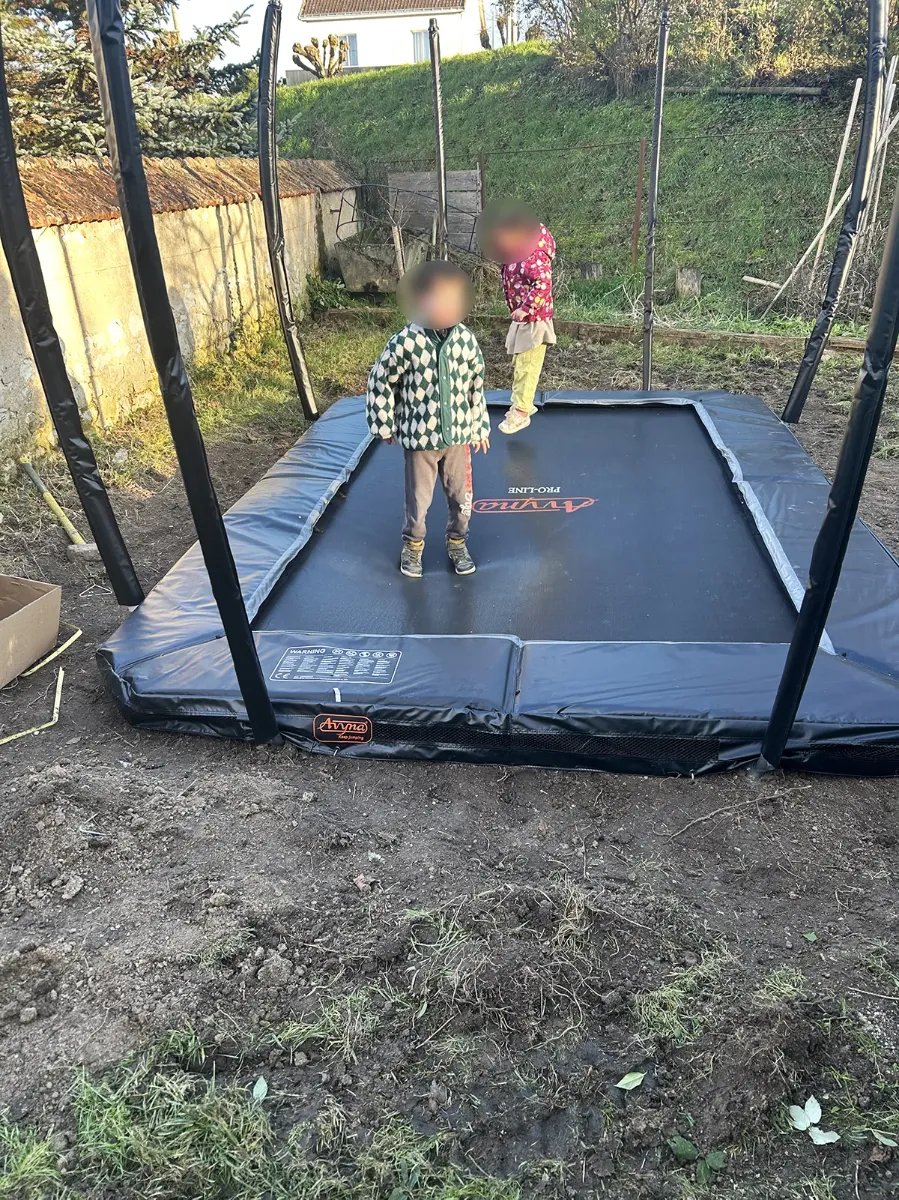 Trampoline enterré au ras du sol dans un jardin clos de murs, filet et poteaux de sécurité en place