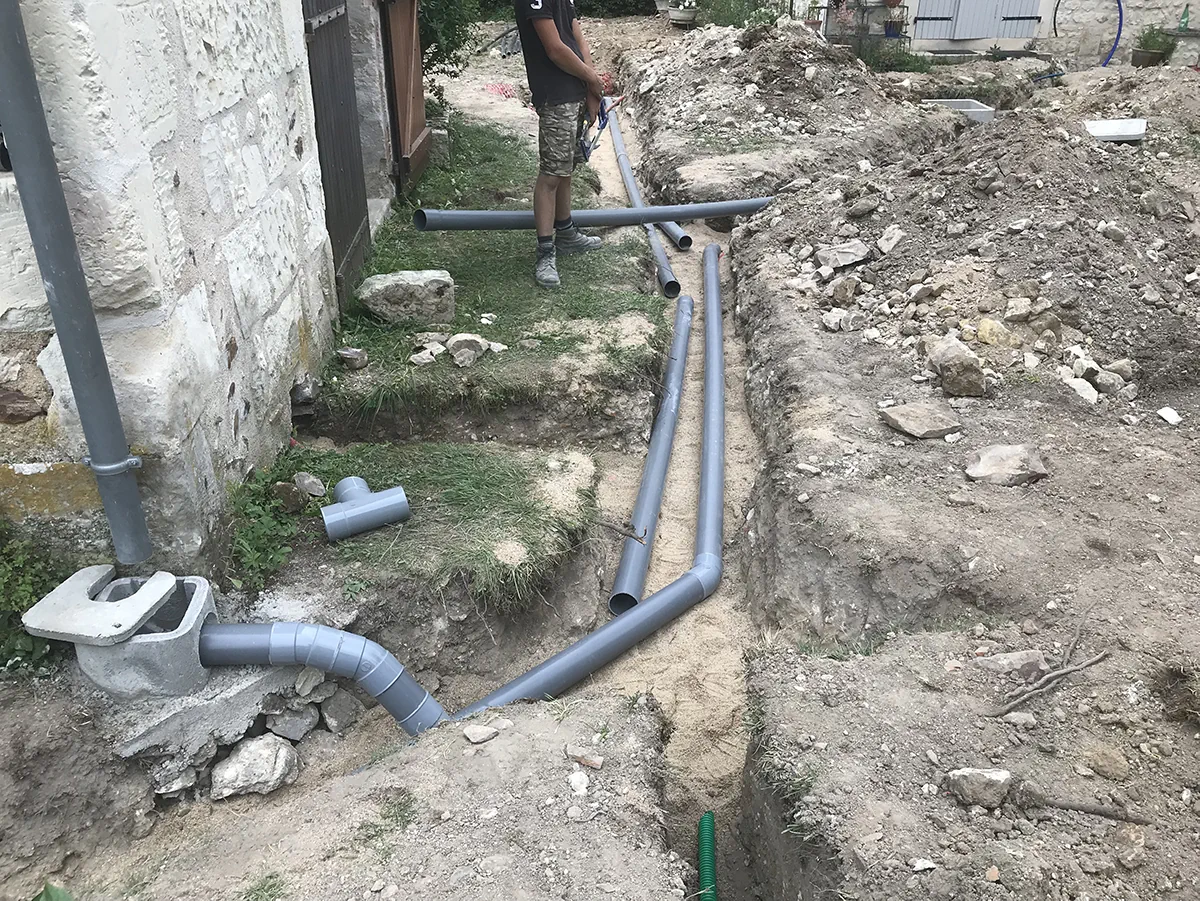 Compagnon assemblant un réseau d'évacuation en PVC dans les tranchées, le long d'une maison de pierre