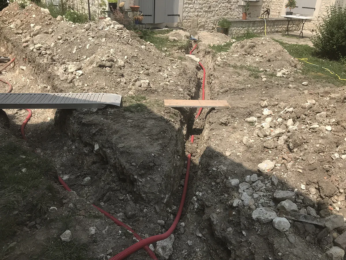 Gaine électrique rouge déroulée au fond d'une tranchée étroite, planche de repère posée en travers
